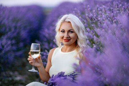 Blonde lavender field holds a glass of white wine in her hands. Happy woman in white dress enjoys lavender field picnic holding a large bouquet of lavender in her hands . Illustrating womans picnic in a lavender field.の写真素材