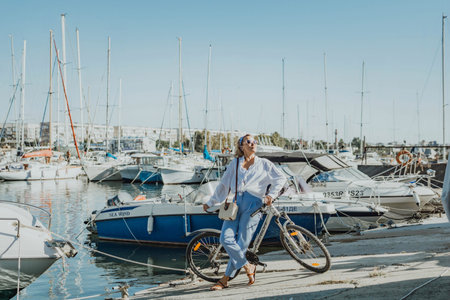 Woman enjoys bike ride along waterfront, Marina surroundings. She is wearing a white shirt and blue jeans, and she has a handbag with her. Capturing outdoor bike ride by waterfront.の写真素材