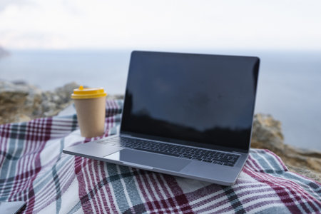 Laptop coffee on blanket with ocean view. Illustrating serene outdoor laptop use. Freelancer enjoying their time outdoors while working or browsing the internet.の写真素材