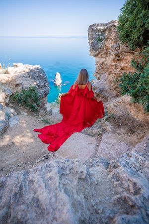 Red dress sea woman. Happy woman with flowing hair in a long flowing red dress stands on a rock near the sea. Travel concept, photo session at seaの写真素材