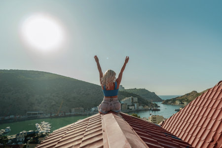 Woman sits on rooftop with outstretched arms, enjoys town view and sea mountains. Peaceful rooftop relaxation. Below her, there is a town with several boats visible in the waterの写真素材