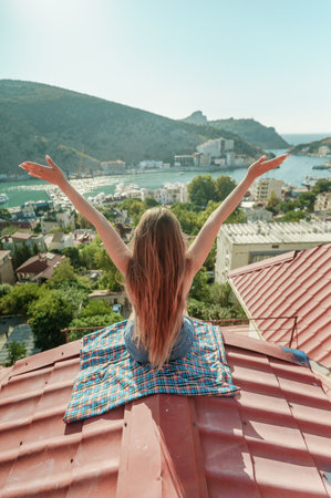 Woman sits on rooftop with outstretched arms, enjoys town view and sea mountains. Peaceful rooftop relaxation. Below her, there is a town with several boats visible in the waterの写真素材