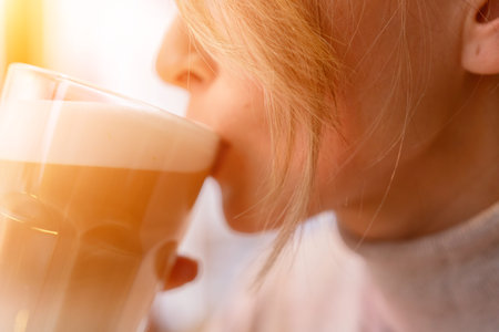 Woman with blonde hair sips cappuccino in a cafe. She is holding the glass up to her face, taking a sip of the drink.の写真素材