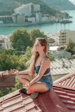 Woman sits on rooftop, enjoys town view and sea mountains. Peaceful rooftop relaxation. Below her, there is a town with several boats visible in the water. Rooftop vantage point.の写真素材