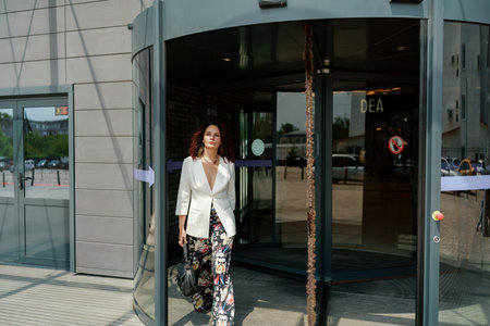 Woman leaves a supermarket. Caucasian model with long dark hair, wearing a white jacket and colored trousers.の写真素材