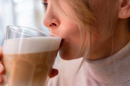 Woman with blonde hair sips cappuccino in a cafe. She is holding the glass up to her face, taking a sip of the drink.の写真素材