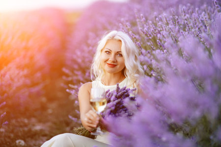 Blonde lavender field holds a glass of white wine in her hands. Happy woman in white dress enjoys lavender field picnic holding a large bouquet of lavender in her hands . Illustrating womans picnic in a lavender field.の写真素材