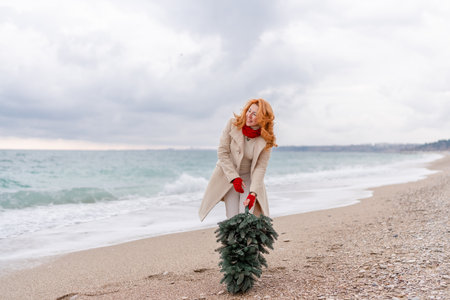 Redhead woman Christmas tree sea. Christmas portrait of a happy redhead woman walking along the beach and holding a Christmas tree in her hands. Dressed in a light coat, white suit and red mittens.の写真素材