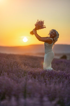 Blonde woman poses in lavender field at sunset. Happy woman in white dress holds lavender bouquet. Aromatherapy concept, lavender oil, photo session in lavenderの写真素材