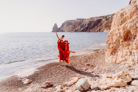 Woman red dress sea. Female dancer in a long red dress posing on a beach with rocks on sunny day....