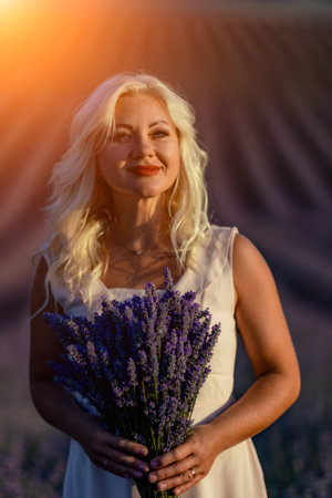 Blonde woman poses in lavender field at sunset. Happy woman in white dress holds lavender bouquet. Aromatherapy concept, lavender oil, photo session in lavenderの写真素材
