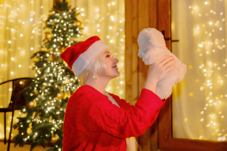 Woman in Santa hat by Christmas tree. Festive setting. Illustrating holiday cheer and celebration. She is posing for a picture, and there are presents nearby, adding to the festive atmosphere.の写真素材