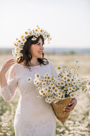 Happy woman in a field of daisies with a wreath of wildflowers on her head. woman in a white dress in a field of white flowers. Charming woman with a bouquet of daisies, tender summer photoの写真素材