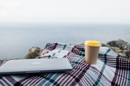 Laptop coffee on blanket with ocean view. Illustrating serene outdoor laptop use. Freelancer enjoying their time outdoors while working or browsing the internet.の写真素材