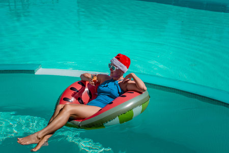 A happy woman in a blue bikini, a red and white Santa hat and sunglasses poses in the pool in an...