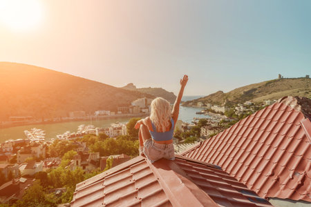 Woman sits on rooftop with outstretched arms, enjoys town view and sea mountains. Peaceful rooftop relaxation. Below her, there is a town with several boats visible in the waterの写真素材