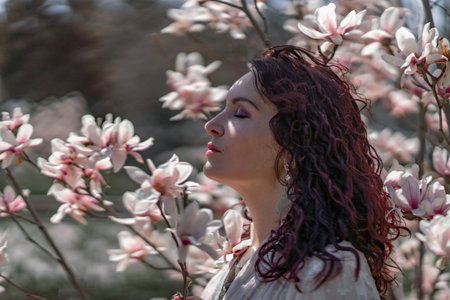 Magnolia park woman. Stylish woman in a hat stands near the magnolia bush in the park. Dressed in white corset pants and posing for the camera.の写真素材
