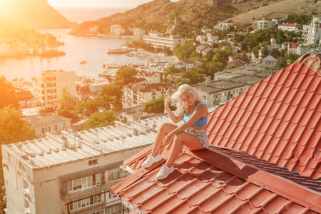 Woman sits on rooftop, enjoys town view and sea mountains. Peaceful rooftop relaxation. Below her, there is a town with several boats visible in the water. Rooftop vantage point.の写真素材