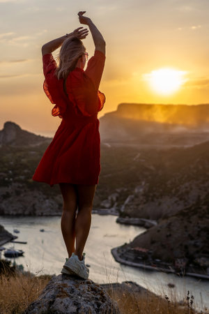 Happy woman standing with her back on the sunset in nature in summer with open hands posing with mountains on sunset, silhouette. Woman in the mountains red dress, eco friendly, summer landscape active restの写真素材