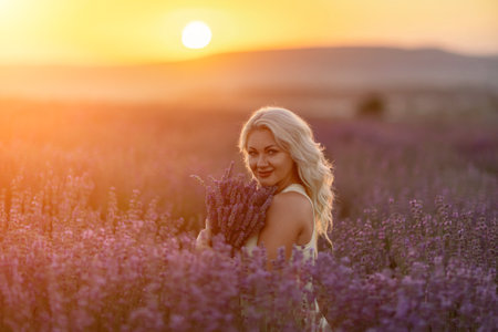 Blonde woman poses in lavender field at sunset. Happy woman in white dress holds lavender bouquet. Aromatherapy concept, lavender oil, photo session in lavenderの写真素材
