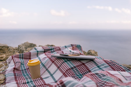 Laptop coffee on blanket with ocean view. Illustrating serene outdoor laptop use. Freelancer enjoying their time outdoors while working or browsing the internet.の写真素材