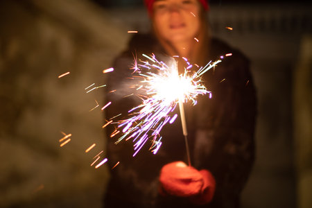 Woman holding sparkler night while celebrating Christmas outside. Dressed in a fur coat and a red headband. Blurred christmas decorations in the background. Selective focusの写真素材