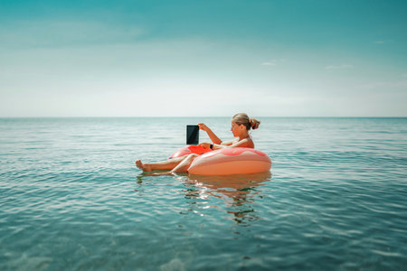 Woman laptop sea. Freelancer woman in sunglases floating on an inflatable big pink donut with a laptop in the sea. People summer vacation rest lifestyle conceptの写真素材