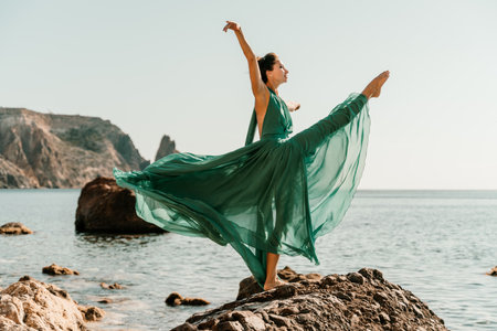 Woman green dress sea. Female dancer in a long mint dress posing on a beach with rocks on sunny day. Girl on the nature on blue sky background.の写真素材