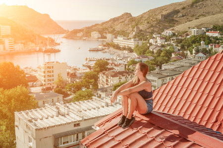 Woman sits on rooftop, enjoys town view and sea mountains. Peaceful rooftop relaxation. Below her, there is a town with several boats visible in the water. Rooftop vantage point.の写真素材