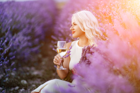 Blonde lavender field holds a glass of white wine in her hands. Happy woman in white dress enjoys lavender field picnic holding a large bouquet of lavender in her hands . Illustrating womans picnic in a lavender field.の写真素材