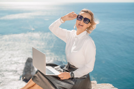 Business woman on nature in white shirt and black skirt. She works with an iPad in the open air with a beautiful view of the sea. The concept of remote work.の写真素材