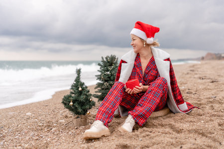 Sea Lady in Santa hat plaid shirt with a red mug in her hands enjoys beach with Christmas tree. Coastal area. Christmas, New Year holidays concepの写真素材