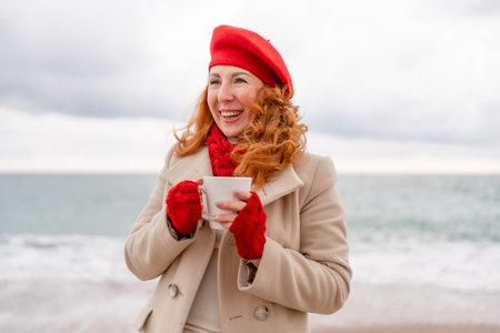 Woman beach in red beret scarf and mitts holding a cup of tea in his hands. Depicting beach relaxation and cozy attire. Walks by the seaの写真素材