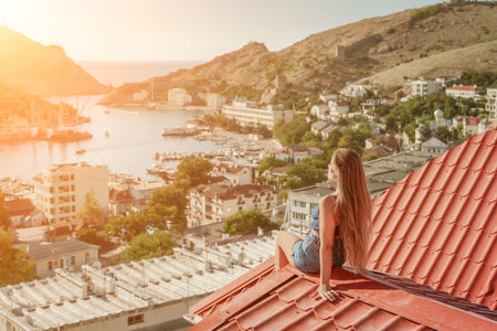 Woman sits on rooftop, enjoys town view and sea mountains. Peaceful rooftop relaxation. Below her, there is a town with several boats visible in the water. Rooftop vantage point.の写真素材