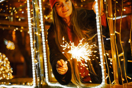 Woman holding sparkler night while celebrating Christmas outside. Dressed in a fur coat and a red headband. Blurred christmas decorations in the background. Selective focusの写真素材