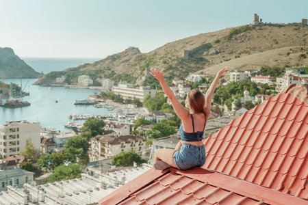 Woman sits on rooftop with outstretched arms, enjoys town view and sea mountains. Peaceful rooftop relaxation. Below her, there is a town with several boats visible in the waterの写真素材