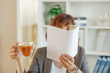 Portrait of a girl holding an open book to her face in one hand and a cup of tea in the other against the background of the interior.の写真素材