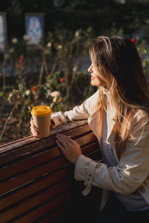 Woman drinks from cup on wooden bench. She is wearing a white shirt enjoying her beverage. The bench...