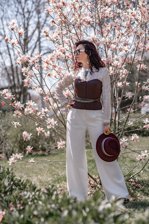 Magnolia park woman. Stylish woman in a hat stands near the magnolia bush in the park. Dressed in white corset pants and posing for the camera.の写真素材