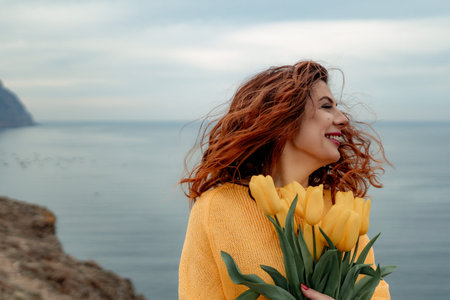 Portrait of a happy woman with hair flying in the wind against the backdrop of mountains and sea. Holding a bouquet of yellow tulips in her hands, wearing a yellow sweaterの写真素材