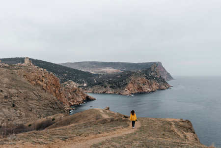Woman stands on cliff overlooking ocean, cloudy day provides soft lighting. Shes wearing a yellow...