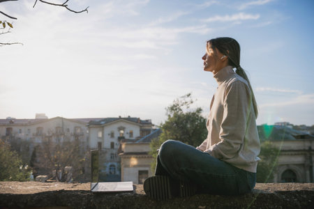 Woman freelancer uses laptop on cement wall outdoors against the sky and the roof of the city. The...