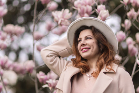 Woman magnolia flowers, surrounded by blossoming trees., hair down, white hat, wearing a light coat. Captured during spring, showcasing natural beauty and seasonal change.の写真素材