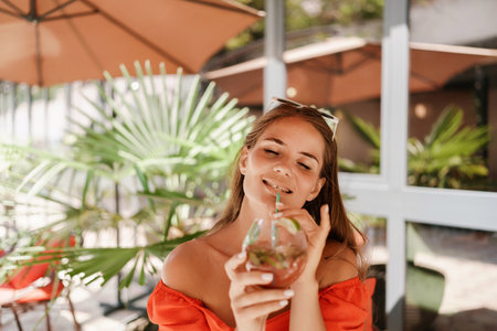 A woman in an orange dress is sitting in a chair with a drink in front of her. She is looking at the camera and she is enjoying her drink. The scene is set in a patio or outdoor area.の写真素材