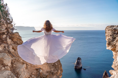 A woman in a white dress stands on a rocky cliff overlooking the ocean. The scene is serene and peaceful, with the womans dress billowing in the wind. The ocean is calm and blue.の写真素材