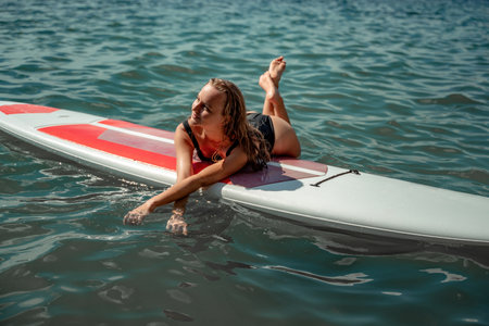 Woman sap sea. Sports girl on a surfboard in the sea on a sunny summer day. In a black bathing suit, he lies on a sap in the sea. Rest on the sea.の写真素材