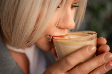 A blonde woman is drinking coffee from a glass. She is smiling and she is enjoying her drink.の写真素材