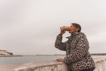 A woman in a leopard print coat is drinking coffee while looking out at the water. The scene is calm...