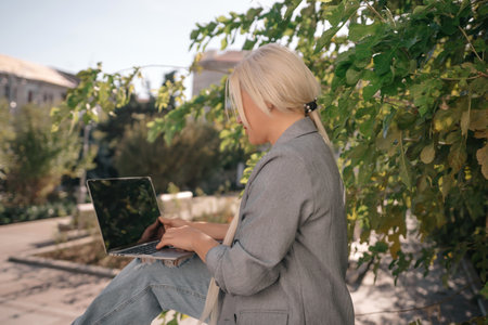 A woman is sitting on a bench with a laptop open in front of her. She is smiling and she is enjoying her time. Concept of relaxation and leisure.の写真素材