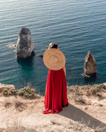 A woman in a red dress stands on a rocky cliff overlooking the ocean. She is wearing a straw hat and she is enjoying the view.の写真素材